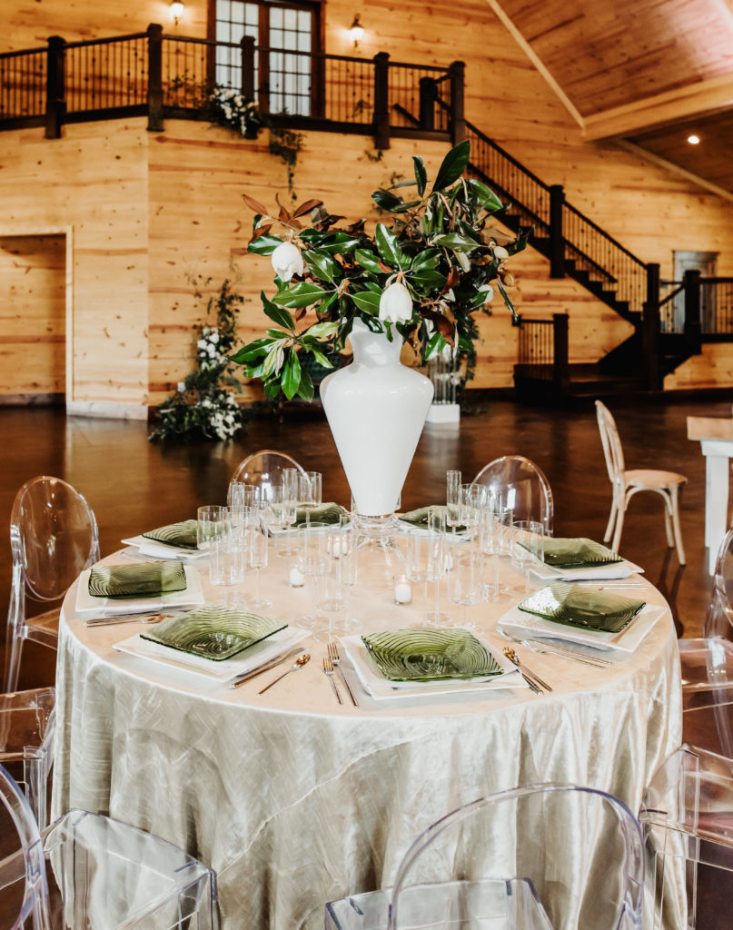Velvet linen clear glass stemware with modern silverware.  ghost chair at round table.  White ceramic vase filled with magnolia leaves and magnolia flowers.  The Carolina Barn at McCormick Farms