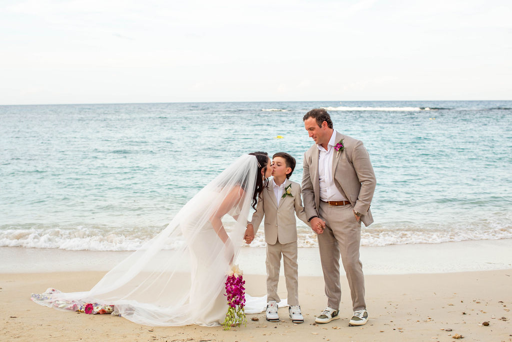 A beautiful beachside vow renewal for couple Devin and Tyler at Sandals Ochi Rios Resort in Jamaica. In this image, the bride and groom pose with their son in front of the ocean.