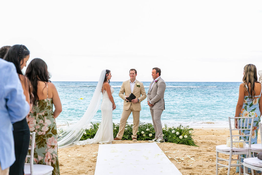 Luxurious vow renewal for couple Devin and Tyler at Sandals Ochi Rios Resort in Jamaica. The bride and groom stand in front of the ocean alongside their officiant while their guests look on.